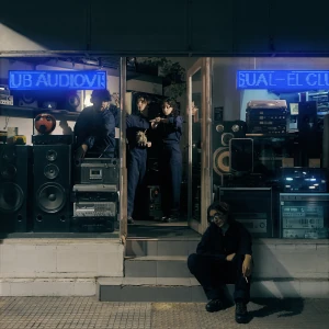 Members of the band El Club Audiovisual posing outside an old retro hi-fidelity music equipment store with LCD signs displaying the bands name